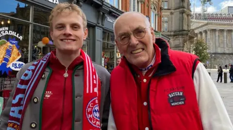 BBC Christian has blonde hair, is wearing a Bayern Munich scarf and a red Bayern jumper. He is standing next to Klaus who is wearing a red gilet and glasses