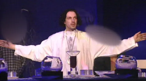 Nine O'Clock Trust Chris Brain leads a service, wearing white robes and with his arms out wide. The lighting is dim with a blue hue.