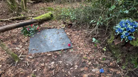BBC/Kevin Reide A gravestone marks a patch of woodland strewn with leaves. Blue roses and re flowers are planted nearby. Holly can also be seen and tangles of shrubbery.