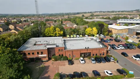 STEVE HUBBARD/BBC An aerial photo of the East of England Ambulance Service Control room and car park. You can see a radio transmitter at the back of the building, along with a row of ambulances, to the front are a number of cars in the car park. The building is red brick and is two stories high to the left hand side with a flat roof, to the right is a hanger for ambulance parking.