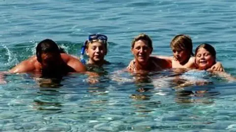 Duncan Ridgley Duncan Ridgley, with his three children, Angus, Claudia, and Sasha, and wife, Penelope, they are all in the sea, you can only see the upper parts of their body, one child is wearing a snorkel and swimming face mask. The sea is deep blue.
