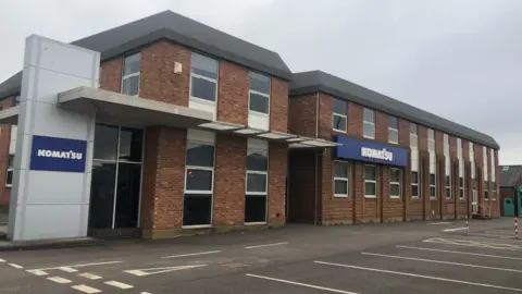 The exterior of the old Meco factory in Worcester. It is a brown brick building with a flat grey roof. Signs on the front and next to the building read "Komatsu". An empty car park is in front of the building.