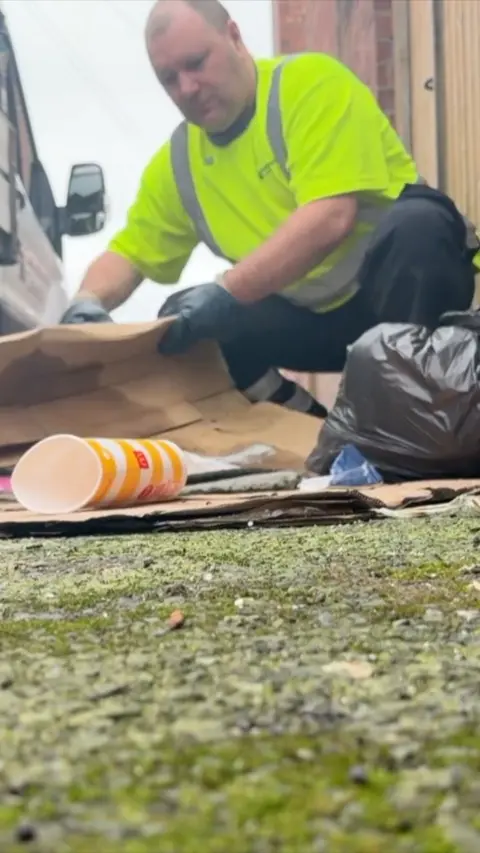 A man in an optic yellow hi-vis t shirt, black trousers and dark blue gloves is crouched down picking up a folded cardboard box. There is a black bag next to his left knee. An empty McDonald's drinking cup is on the left. Mossy pavement takes up a third of the frame from the bottom. 