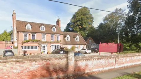 Google A Google Maps Streetview image of The Lodge care home. On the right of the image is a red sign which has The Lodge written on it in gold writing. On the left of the image is a large house-like building, in which the care home is located. There are cars parked outside.