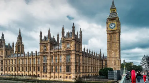 General view of the Houses of Parliament in London, with clouds and grey skies overhead. Palace of Westminster and Big Ben clock face.