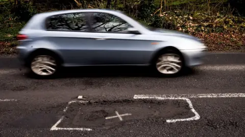 PA Media A light blue car driving past a pothole, which has been marked with paint.