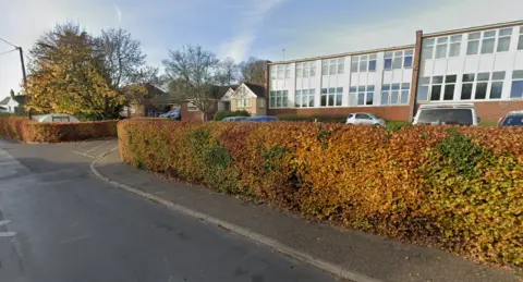 Google A 1970s-style school building with a bank of white-framed windows and flat roof, viewed from a roadside with head-height hedging in between.