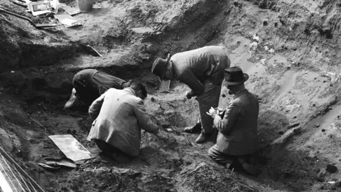 Trustees of the British Museum/PA A black and white image of archaeologists in the 1930s uncovering a Anglo-Saxon burial ship in the ground. Four people look at the ground as one of moves their hand over the earth.