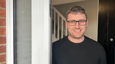 A man smiling gingerly at the open door of his house