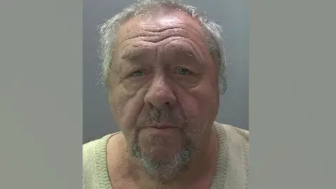 Gheorghe Hirtopeanu, who has short grey hair and a grey goatee-style beard, wearing a yellow v-neck top. He is looking at the camera in a police mug shot.