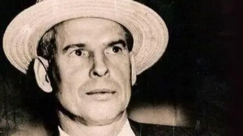 Llewellyn Morris Humphreys - better known as Murray the Hump - is smartly dressed in collar and tie, he is wearing a cream colour hat with a black ribbon around it. He looks every inch a society gentleman