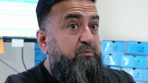 Instagram An Asian man in his 50s, with a long black beard streaked with grey and black hair, speaks into the camera while wearing a black dental uniform