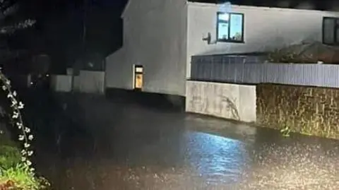 Axminster Fire Station A flooded road next to a white house during the night time as heavy rain falls. A light is on in a window on the ground floor of the property and another is on in a larger window on the top floor.