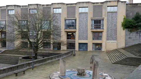 Winchester Crown Court, a plain building resembling an office block, sits beyond a courtyard with a bare tree and an art feature with three small wooden pillars on a mosaic. To the right, a set of long steps ascend until they disappear out of shot. 
