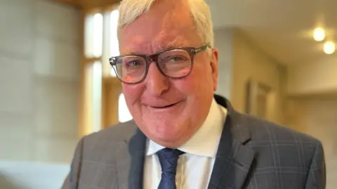 BBC Fergus Ewing has short white hair. He is wearing glasses and a grey suit over a white shirt with a blue tie. Is pictured indoors.