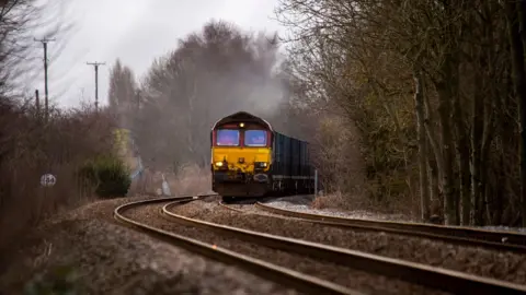 A cargo train approaching in the distance on a track. It has a yellow cab with a red top. There is mist coming off the roof. Trees line the track.