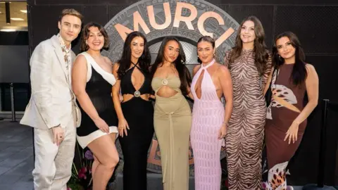 MURCI A man on the left stands next to six women in front of a sign saying 'MURCI' . He wears a cream linen suit, while the women all wear maxi dresses of different colours and patterns. 