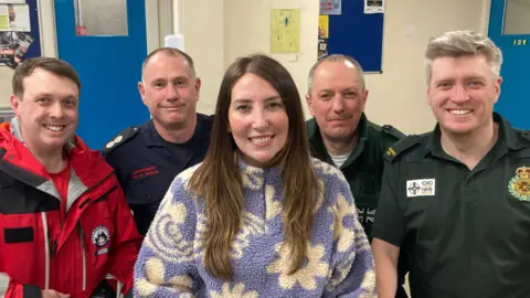 BBC A group photo showing a woman in the centre with long brown hair wearing a purple and white fleece with flowers on it. Four men surround her wearing different uniform - one is in a red and black jacket, another in a blue shirt, and two on the right are wearing dark green shirts.