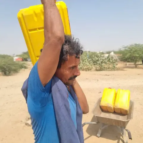 A man in a blue T-shirt carries a large yellow canister on his shoulder. There are two more canisters in a wheelbarrow behind him.