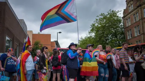 Newcastle Pride 2024. A crowd of people line the street. Many are holding rainbow flags and are dressed in colours associated with Pride.