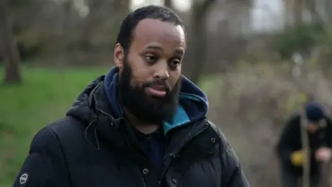 Head-and-shoulders image of a Mahamed Hashi, seen in a dark jacket in a park, with another person planting a young tree in the background.