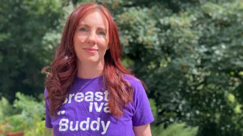 Jennifer has dyed red hair that falls into curls. She is wearing a purple t-shirt that reads: 'Breadtival Buddy'.