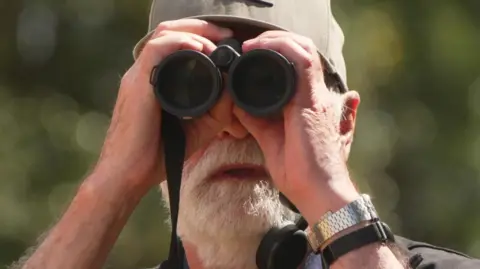 Nigel Hewitt is holding a black pair of  binoculars and wearing a cap. He has a white beard.