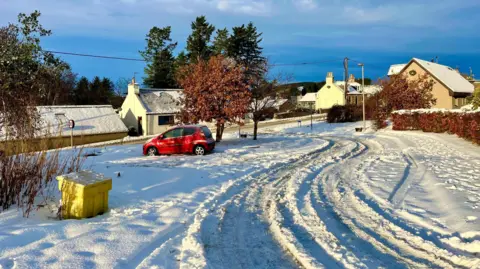 There are tyre tracks through snow at the village of Auchleven. There are a handful of houses among trees with snow on their roofs. There is a yellow plastic grit box and a little red car parked next to a tree.