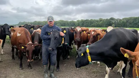 Farmer Tom Pemberton stands in a field of cows. They are surrounding him and he is stroking one of them by its ears.