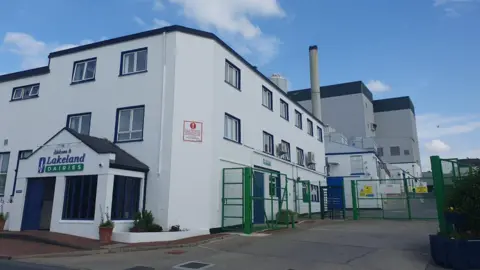 A factory building painted white, with a number of gates and security fences. A sign saying lakeland Dairies is above a blue door at the front of the building.