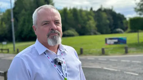 A man with white hair and a white goatee and a white shirt with a UCU lanyard round his neck. He is standing in front of a green grassy area with trees in the background.