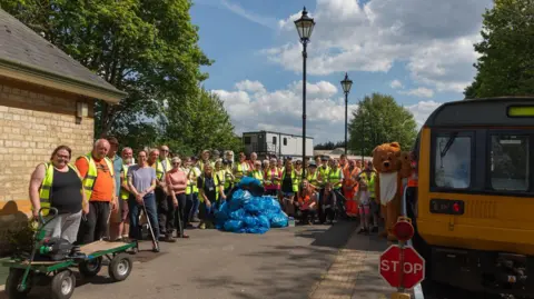 The Auckland Project About 50 volunteers in high visibility vests are standing on the platform a Stanhope station and smiling. There is a pile of blue waste bags in front of them. A yellow train is on the rails. 