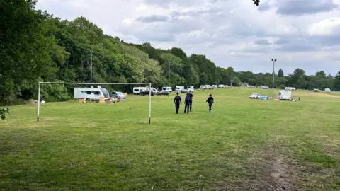 BBC/Sara Thornton Four people officers stand at the entrance to a park, with a metal gate across the front and a sign saying Waterer's Park, with a row of caravans along the edge of the field.