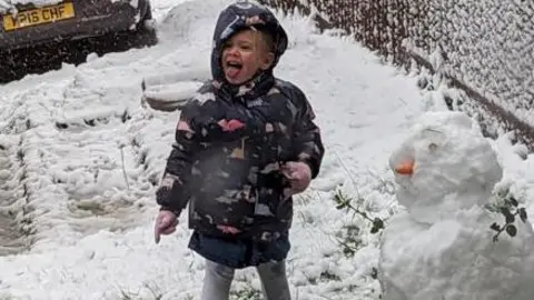 Chloe Thompson A young girl in boots and anorak stands in snow with a snowman.