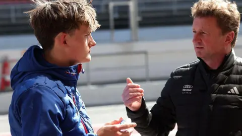 Getty Images Allan McNish in a dark Audi jacket is deep in conversation with young racer Freddie Slater in a blue jacket with light brown hair. They are at a race track.