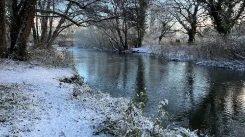 Shaun Whitmore/BBC A dusting of snow on the banks of a river. There are trees in each bank and their branches have snow on them. The trees are reflected in the water.