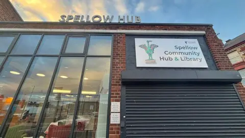 A brick building with large glass windows and metal shutters with a sign saying, 'Spellow Hub' against a sunny sky 