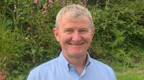 Steve Rubidge is smiling at the camera as he stand in a garden. He has short grey hair and is wearing a pale blue shirt. There are ferns and pink flowers in the hedging behind him.
