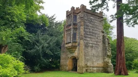 A crenelated stone tower with a large arched doorway. Above the door are two ornate bay windows, one above the other. The tower has broken stones on one side suggesting it was part of a larger building. It is surrounded by trees.