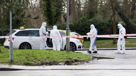 Pacemaker Four forensic officers wearing white body suits and face masks investigate a white saloon taxi car that is parked in an empty car park. The photo is taken from a distance. There is police tape cordoning off the area.