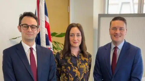 Josh Dean A line of three people (left to right) - Josh Dean with short hair and black-framed glasses, wearing a white shirt, red tie and grey jacket, Rachel with long dark hair wearing a brown and yellow top, and Wes Streeting with short dark hair, wearing a pale blue shirt and red tie with a blue jacket. There is a Union flag behind them, along with a yellow and green patterned wall and a whiteboard.