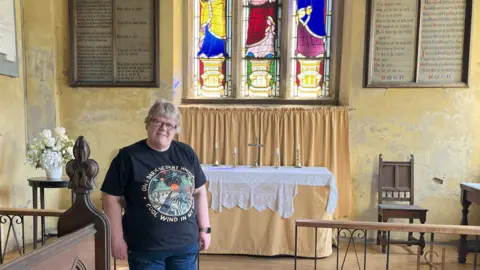 A middle-aged woman wearing a black t-shirt, she has short cropped fair hair and wears glasses. She stands at the front of the church, where an alter is placed and a stained glass window can be seen in the background.