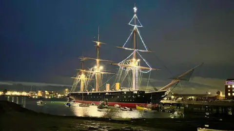 HMS Warrior at night in Portsmouth harbour. The mast is lit up with Christmas lights in the shape of a Christmas tree.