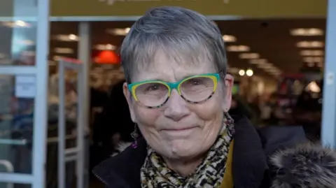 Anne Kingsley is smiling at the camera while wearing a coat and a scarf. She has green glasses and short grey hair. 