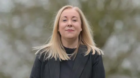 Ulster Wildlife Dawn Miskelly, a woman with long, straight, blonde hair, smiles at the camera. She is wearing black pinstriped suit jacket over a black top. A tree in the background is blurred out. 