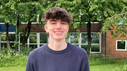 A young man with brown hair and braces wears a navy jumper and smiles in front of trees and a building