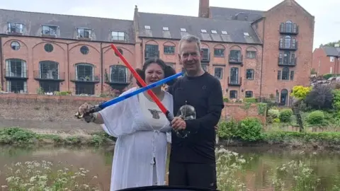 Jo Yarnall A woman dressed as Princess Leia in a white flowing top, with her hair in two big buns at the side of her head, holding a blue toy lightsaber, which is crossed with a red lightsaber held by a man next to her in black clothing. They stand in front of the river and a row of red brick buildings