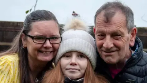 Sam, Poppi and Ian Frisby on a grey day. They are all smiling at the camera. Sam has long dark hair and wears a yellow dress with glasses. Poppi has long auburn hair and wears a white bobble hat. Ian has grey hair and wears a black jacket.