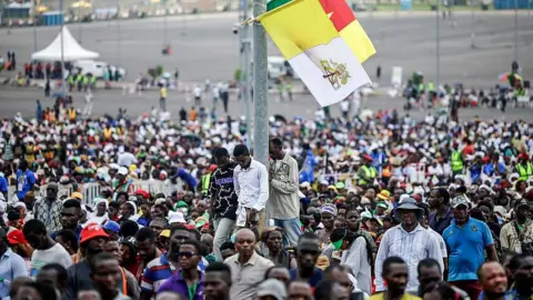 AFP via Getty Images Crowd of people with a pole in the centre with three men stood against it. the pole has a Vatican flag and a Cameroon flag attached. They all look to be listening