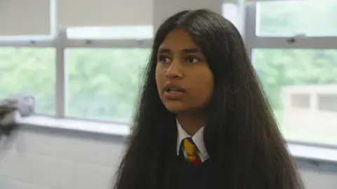 A girl in a school uniform with a yellow, red and blue tie and black sweater sat in a classroom